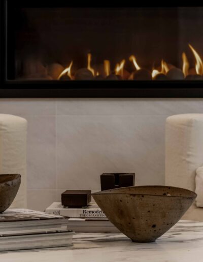 A cozy living room scene with a lit fireplace. Two ceramic bowls rest on a stack of design books on a white marble table.