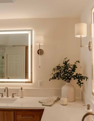 Modern bathroom with a lit square mirror above a sink, flanked by two wall sconces. A vase with greenery and a soap dispenser sit on the countertop.