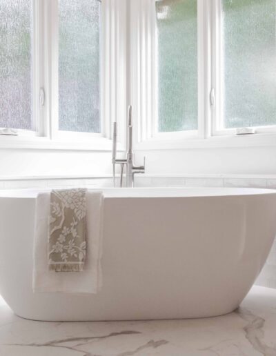 A white freestanding bathtub with a floral towel draped over the side is positioned in front of frosted windows, in a well-lit bathroom with a marble floor.