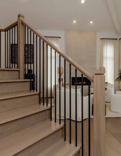 Spacious living room with large windows, light wood flooring, and a staircase with black railings. Features a white sofa, fireplace, and indoor plants.