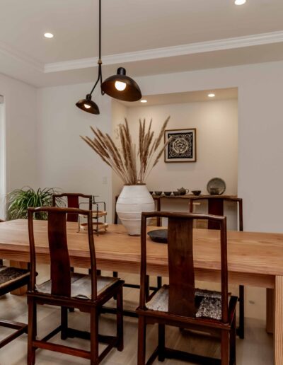 A modern dining room with a wooden table, six chairs, pendant lights, a large vase with dried plants, and a window showing greenery outside.