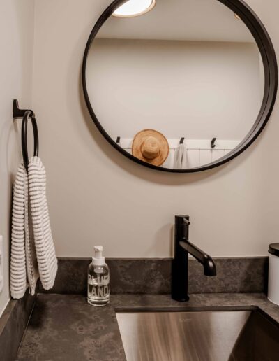 Bathroom with a round mirror, black faucet, stone countertop, sink, two white towels on a hook, soap dispenser, and a reflection of a hat.