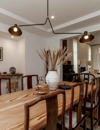 Dining room with a wooden table, chairs, and decorative vase. Modern lighting hangs above. In the background, a living room with a large window and trees outside.