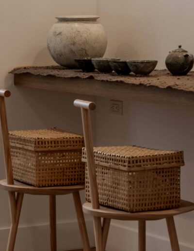 Two woven baskets on wooden chairs, with a rustic shelf above holding a large ceramic vase and several smaller pottery items against a plain wall.