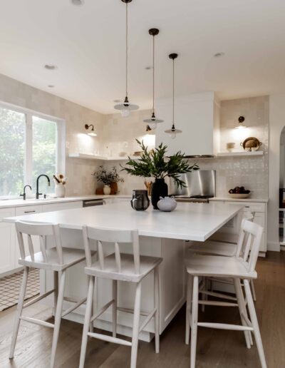 Modern kitchen with a large central island, white cabinetry, pendant lights, and bar stools. Natural light enters through a window above the sink.