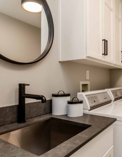 Modern laundry room with a black countertop, sink, and faucet. Two white storage jars sit next to the sink. A washer and dryer are positioned next to white cabinets and a round mirror.