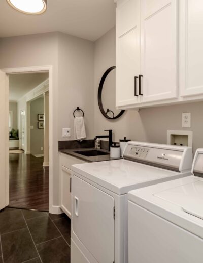 Modern laundry room with white washer and dryer, white cabinets, and black accents. Open door leads to a hallway with wooden flooring.