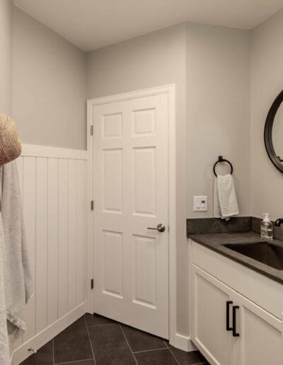 Laundry room with a white door, round mirror, black faucet, and white cabinets. A beige hat and towels hang on the wall. Gray tile floor and countertop.