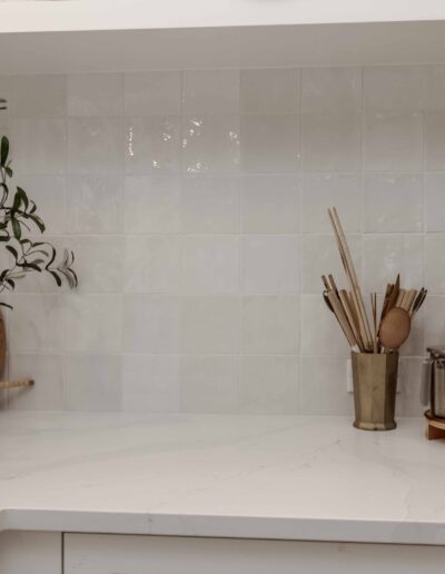 A white kitchen counter with a potted plant, cutting board, and containers holding utensils against a tiled backsplash.