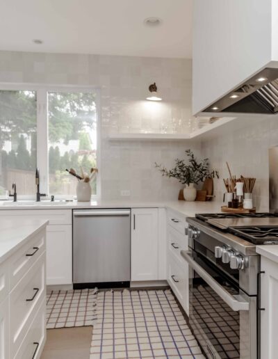 A modern kitchen with white cabinets, stainless steel appliances, and a tiled floor. A window over the sink provides natural light. Decorative plants and kitchenware are on the counters and shelves.