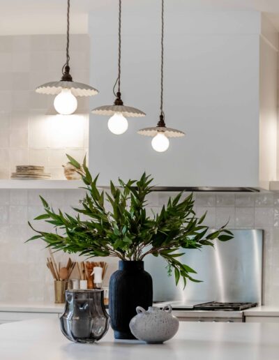 Modern kitchen with three pendant lights above a white island, featuring a black vase with green leaves and a gray decorative bowl. Stainless steel backsplash and neutral tones in the background.