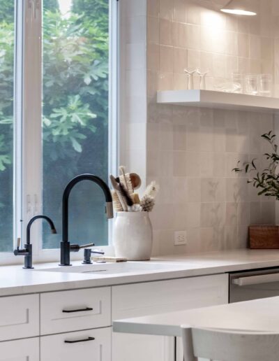 A bright kitchen with white cabinets, a black faucet, open shelves with dishes, and plants on the counter. A large window provides natural light.