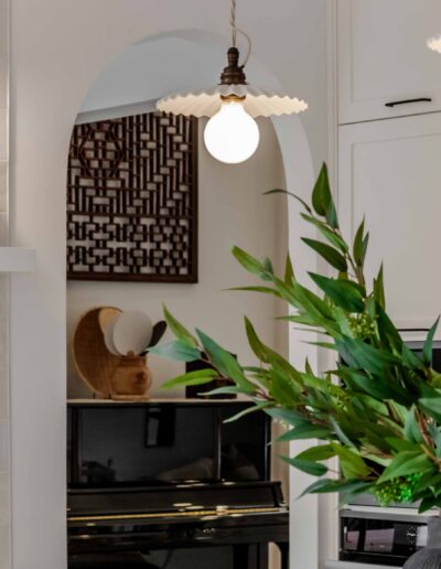 A kitchen with modern pendant lights, a white cabinet, and stainless steel refrigerator. A vase with green foliage is on the counter, and decorative items on a shelf.