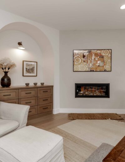 A cozy living room with a white armchair, woven chair, and a wooden console. The room features a framed artwork, a wall-mounted fireplace, and decorative items.