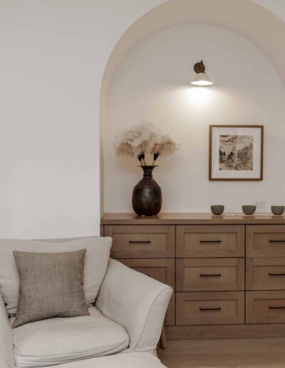 A cozy nook with a cushioned armchair, a wooden cabinet with drawers, a vase of dried flowers, wall art, and a small lamp under an arched alcove.