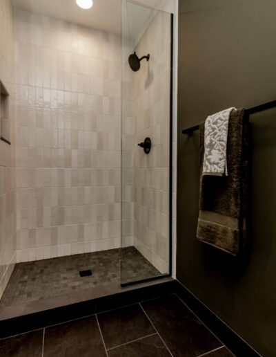 Bathroom with a walk-in shower featuring white tiled walls, dark floor tiles, and a glass door. A towel hangs on a black rod adjacent to the shower.