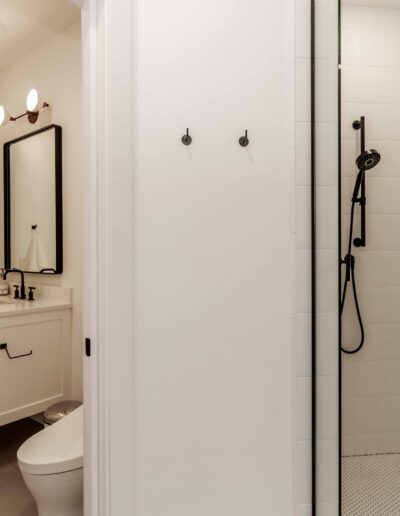 Modern bathroom with a white vanity, mirror, and wall-mounted light above. A walk-in shower with a black handheld showerhead, grab bar, and two hooks is on the right.