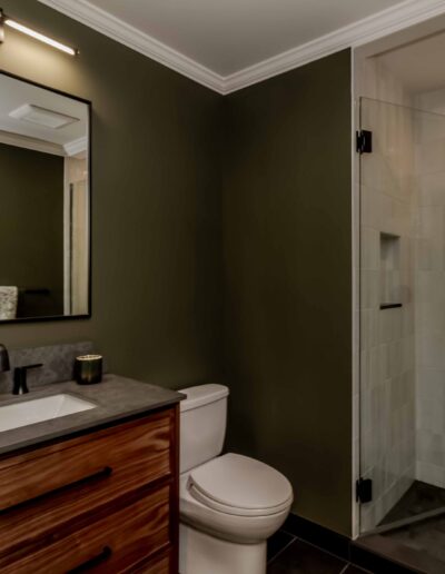 Modern bathroom with dark green walls, a wooden vanity with a black faucet, a rectangular mirror, a toilet, and a glass-enclosed shower.