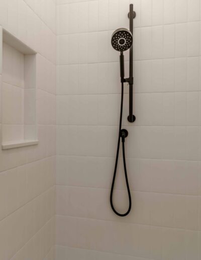 A white tiled shower with a black handheld showerhead, a control knob, a grab bar, and a small recessed shelf.