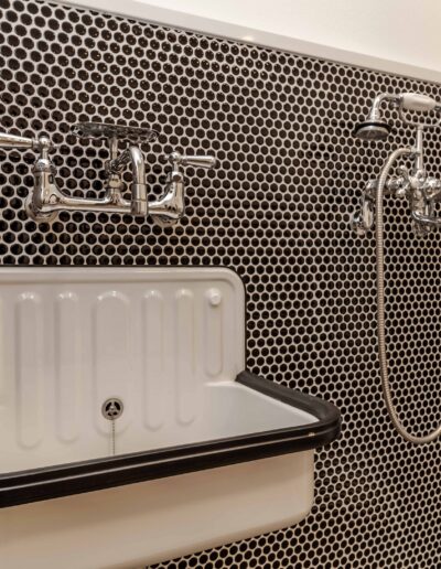 A retro-style bathroom with a white wall-mounted sink. The wall features small black hexagonal tiles and a dual-handle faucet. A shower area with silver fixtures is on the right.