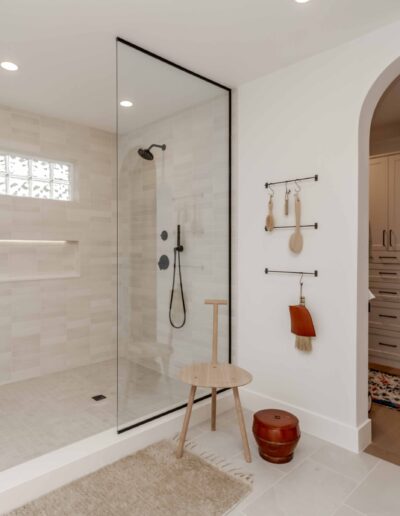 Modern bathroom with glass shower, light tile, a wooden stool, and a doorway leading to a closet with drawers. Rugs and hanging utensils are visible near the entrance.