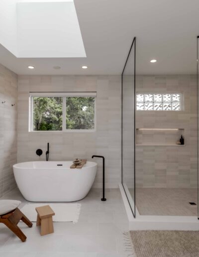 Modern bathroom with freestanding tub, glass-enclosed shower, and a wall-mounted fireplace. There's a wooden chair and stool on a tiled floor, under a skylight and a large window.