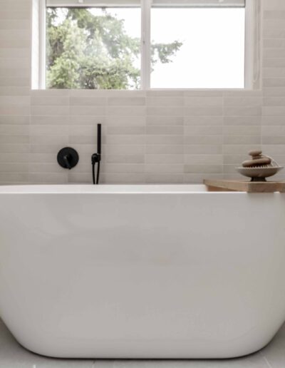 Minimalist bathroom with a white freestanding bathtub, black fixtures, and a window. A wooden board with stacked stones rests on the tub's edge.