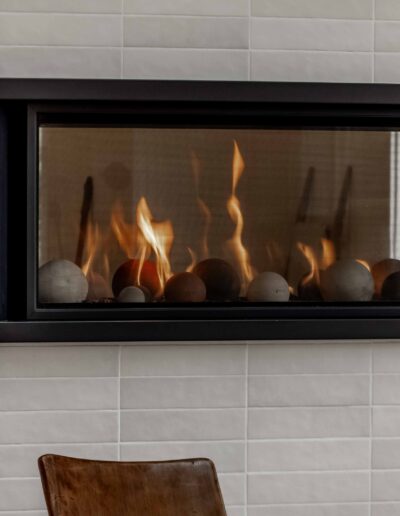 A modern gas fireplace with visible flames is set in a tiled wall, with a white towel hanging on the left and a wooden chair partially visible in the foreground.