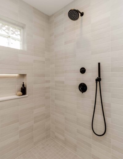 A modern shower with beige tile walls and floor, featuring a black rain showerhead, handheld shower, and built-in shelf with toiletries.