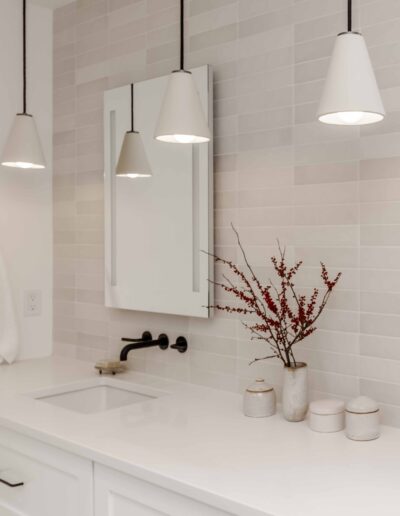 Modern bathroom with white countertop, wall-mounted mirror, three pendant lights, and a vase with red branches.