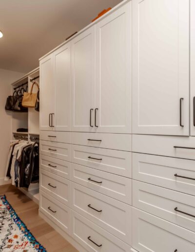 A spacious, modern walk-in closet with white cabinets, hanging clothes, handbags, a colorful rug, and a full-length mirror.