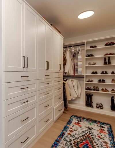 A walk-in closet with white cabinets, drawers, and shelves. Clothes hang on rods, and shoes are neatly arranged. A colorful rug lies on the wooden floor.