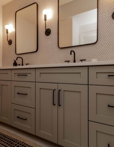 Modern bathroom with dual sinks, hexagonal tile backsplash, two mirrors, wall-mounted lights, and a gray vanity with black hardware. A towel hangs on the wall.