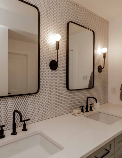 A modern bathroom with a double vanity, two rectangular mirrors, black faucets, and a hexagonal tile backsplash. A towel hangs on the wall, and a soap dispenser is on the counter.