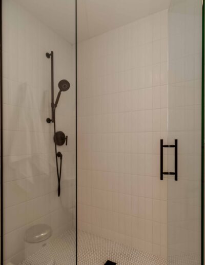 A modern shower with a glass door, white tiled walls, black shower fixture, and a black handle. There's a towel hanging inside.